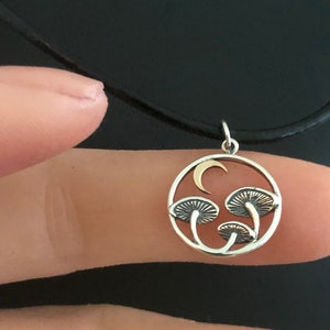 May include: A silver necklace with a round pendant featuring a crescent moon and three stylized mushrooms. The pendant is made of silver and gold.