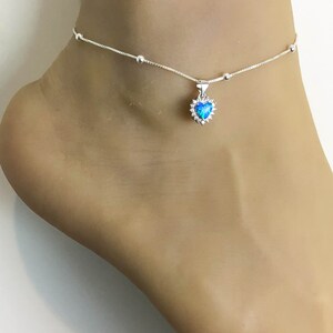 Könnte beinhalten: Silbernes Fußkettchen mit einem kleinen Herz-Anhänger mit einem blauen Opal-Stein, umgeben von klaren Zirkonia-Steinen.