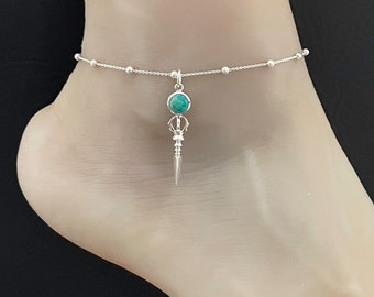 May include: Silver anklet with a turquoise gemstone pendant. The pendant is shaped like a stylized dagger with a pointed tip.
