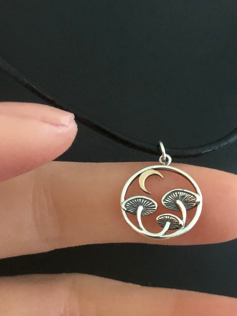 May include: A silver necklace with a round pendant featuring a crescent moon and three stylized mushrooms. The pendant is made of silver and gold.