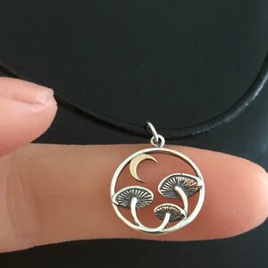 May include: A silver necklace with a round pendant featuring three gold mushrooms and a crescent moon. The pendant is on a black cord.