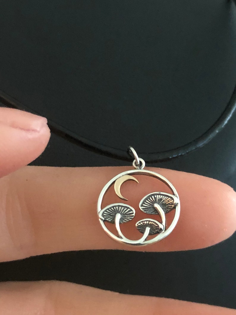 May include: A silver necklace with a round pendant featuring three gold mushrooms and a crescent moon. The pendant is on a black cord.