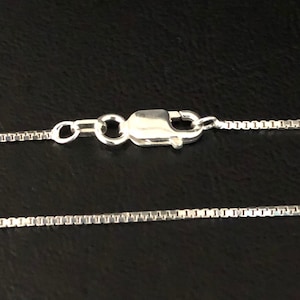 May include: A close-up of a silver box chain necklace with a lobster clasp. The chain is made of small, square links. The clasp is attached to the chain with a small jump ring.