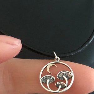 May include: A silver necklace with a circular pendant featuring a crescent moon and three stylized mushrooms. The pendant is on a black cord.