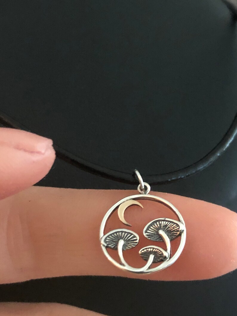 May include: A silver necklace with a circular pendant featuring a crescent moon and three stylized mushrooms. The pendant is on a black cord.