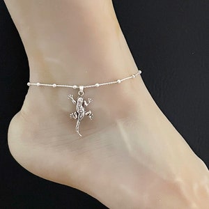 May include: A silver ankle bracelet with a small silver lizard charm. The bracelet is made of a delicate chain with small beads.