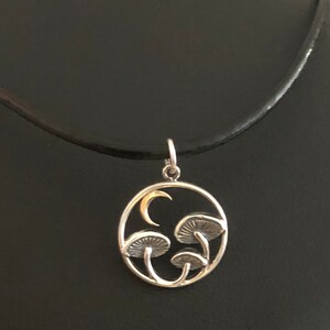 May include: A silver necklace with a circular pendant featuring a crescent moon and three stylized mushrooms. The pendant is on a black leather cord.