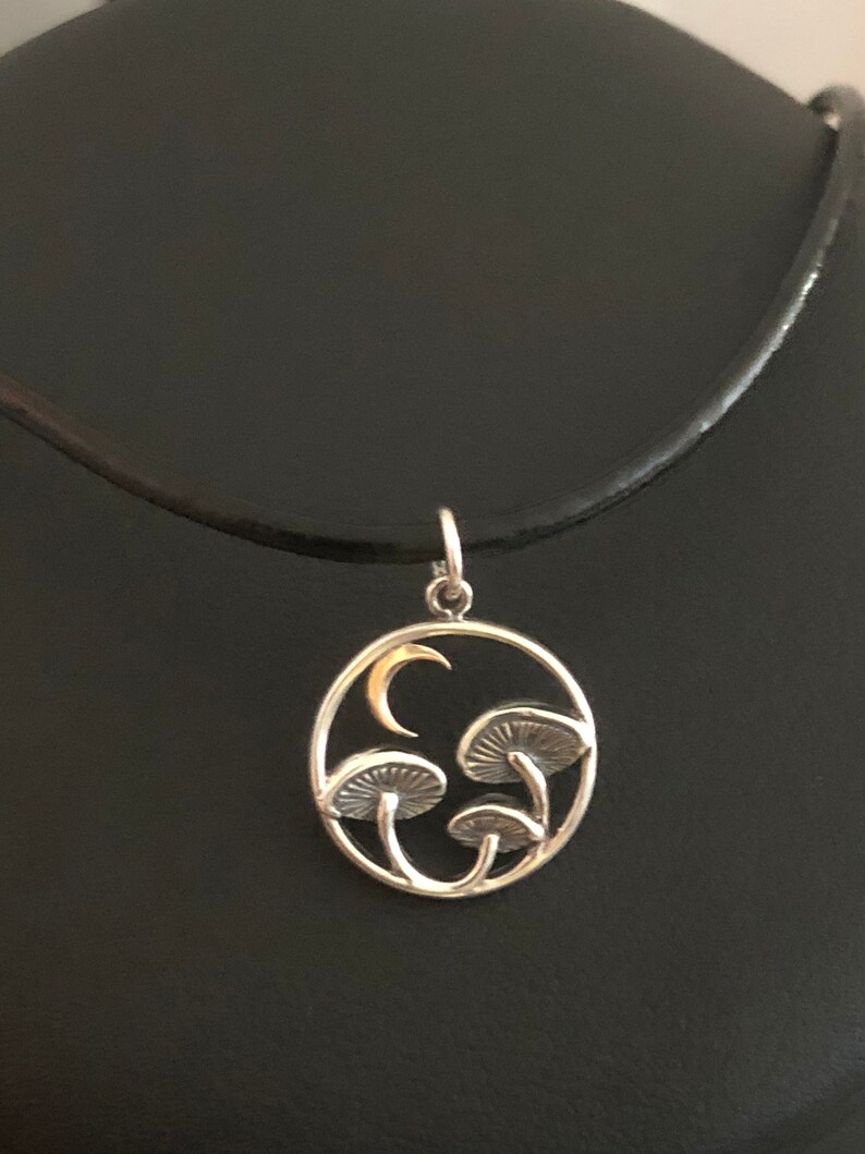 May include: A silver necklace with a circular pendant featuring a crescent moon and three stylized mushrooms. The pendant is on a black leather cord.