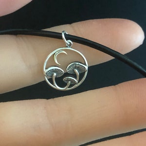 May include: A silver necklace with a circular pendant featuring a crescent moon and three stylized mushrooms. The pendant is on a black cord.