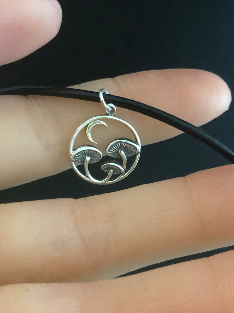 May include: A silver necklace with a circular pendant featuring a crescent moon and three stylized mushrooms. The pendant is on a black cord.