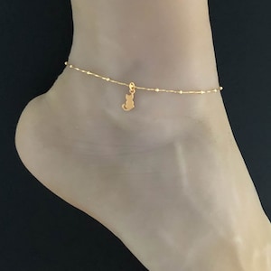 Puede incluir: Un tobillo de oro con un pequeño dije de gato. El tobillo está hecho de una cadena delicada con pequeñas cuentas.
