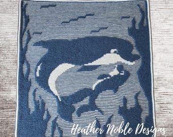 May include: A square crochet blanket featuring a dark blue and white graphic design of two dolphins swimming among seaweed. The pattern is created with a striped texture throughout the piece. The text Heather Noble Designs is printed in white at the bottom right corner of the blanket.
