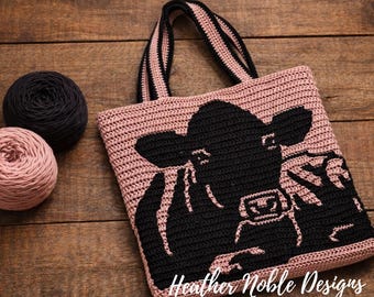 May include: A dusty pink crochet tote bag featuring a black cow graphic on the front and two black and pink striped handles. The text Heather Noble Designs is printed in white script at the bottom right corner of the bag.