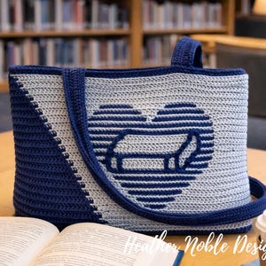 May include: A handmade crochet tote bag in shades of blue and gray, featuring a heart-shaped design with a dachshund silhouette. The bag has a shoulder strap and a top handle. The text "Heather Noble Designs" is visible.