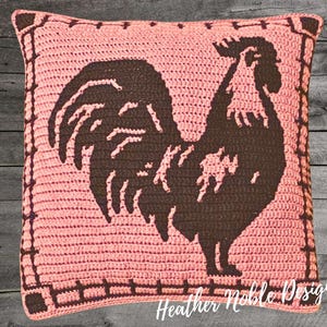 May include: A square, crocheted pillow in a light pink hue, featuring a dark brown rooster silhouette. The rooster is centered on the pillow, with a decorative border. The text "Heather Noble Designs" is visible at the bottom.