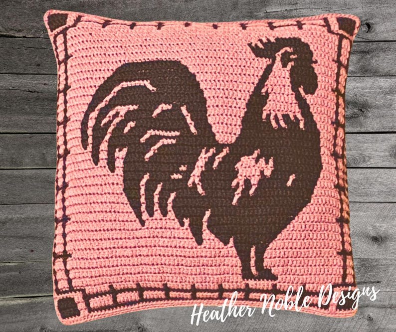 May include: A square, crocheted pillow in a light pink hue, featuring a dark brown rooster silhouette. The rooster is centered on the pillow, with a decorative border. The text "Heather Noble Designs" is visible at the bottom.