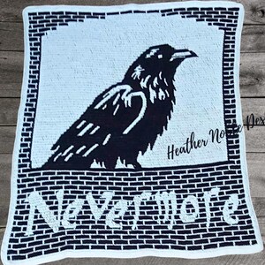 May include: A black and white crochet blanket featuring a raven design. The raven is perched above the word "Nevermore" in white block letters. The blanket has a brick-like border and the text "Heather Noble Designs".