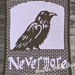 May include: A crocheted blanket featuring a raven design in shades of brown and white. The raven is set against a white background, framed by a brick-like border. The words "Nevermore" are displayed at the bottom.