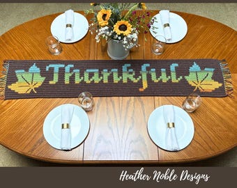 Puede incluir: Un camino de mesa rectangular de ganchillo marrón con la palabra Thankful en un degradado de azul, verde y amarillo. El diseño incluye dos grandes motivos de hojas de arce en los extremos, cada uno con el mismo degradado de color.