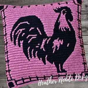 May include: A square, crocheted pillow in shades of pink and black. The pillow features a black silhouette of a rooster in the center. The border is a darker shade of pink with a decorative stitch. The text "Heather Noble Designs" is at the bottom.