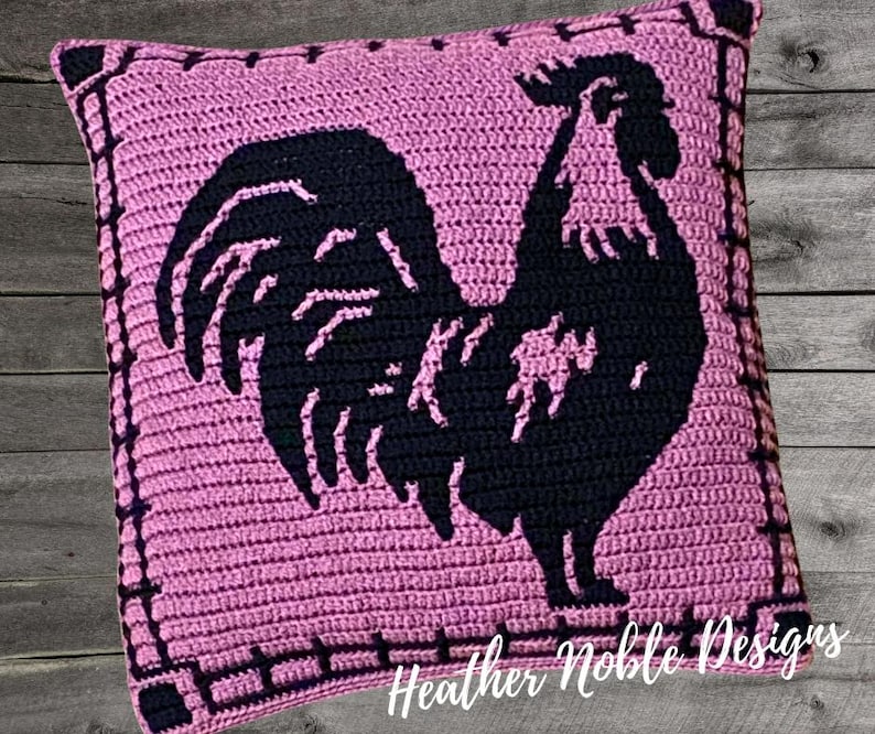 May include: A square, crocheted pillow in shades of pink and black. The pillow features a black silhouette of a rooster in the center. The border is a darker shade of pink with a decorative stitch. The text "Heather Noble Designs" is at the bottom.