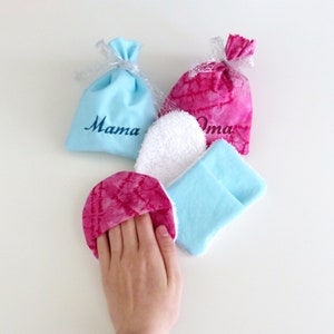 Pode incluir: Duas pequenas sacolas de presente de tecido, uma azul com a palavra "Mama" bordada e outra rosa com a palavra "Oma" bordada. Há também três panos pequenos, um branco, um azul claro e um rosa.