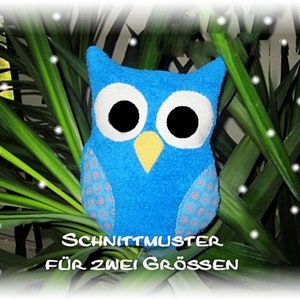 Puede incluir: Un juguete de peluche azul en forma de búho con ojos blancos y un pico amarillo. El búho está sentado frente a una planta verde. El texto "Schnittmuster für zwei Grössen" está escrito debajo del búho.