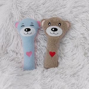 以下が含まれることがあります： 2つのテディベア型のぬいぐるみのおもちゃ。1つはピンクのハートと耳が付いた水色、もう1つは赤いハートと耳が付いたタン色です。どちらも顔が刺繍されており、ふわふわの白い表面にあります。