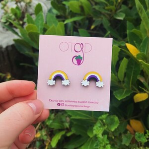 May include: A pair of rainbow-coloured wooden earrings with a purple, white, yellow, and light blue design. The earrings are crafted with sustainable bamboo plywood and are displayed on a pink card with the text "OTGD" and "@onthegrapevinedesign".