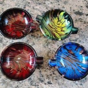May include: Four round ashtrays with a marbled glass design. The ashtrays are in shades of red, green, yellow, and blue. Each ashtray has a small handle. The ashtrays are on a textured surface.