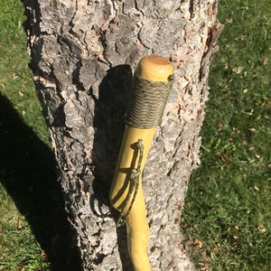 May include: A wooden walking stick with a brown leather wrap and a green camouflage strap. The stick is leaning against a tree trunk.