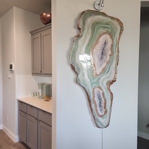 May include: A large, green and white agate geode wall art piece with gold accents. The geode is lit from within, creating a warm glow. The geode is mounted on a white wall.