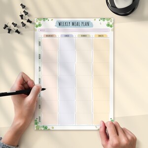 May include: A weekly meal plan with pastel-colored boxes for each meal of the day. The title "WEEKLY MEAL PLAN" is in a blue banner. Green leaf illustrations decorate the top and bottom. A hand is holding a black pen, ready to write.