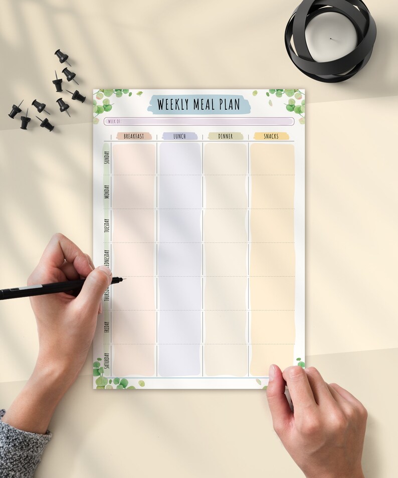 May include: A weekly meal plan with pastel-colored boxes for each meal of the day. The title "WEEKLY MEAL PLAN" is in a blue banner. Green leaf illustrations decorate the top and bottom. A hand is holding a black pen, ready to write.