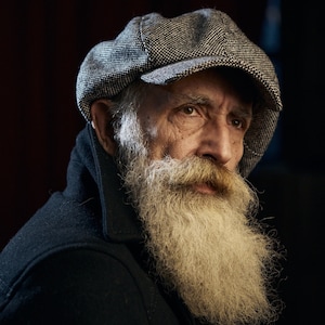 May include: A close-up portrait of an older man with a long white beard. He is wearing a black jacket and a grey newsboy cap. The man is looking off to the side.