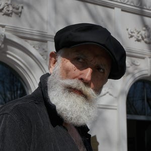 May include: A black corduroy jacket with two chest pockets and a black beret. The jacket is buttoned and has a vertical ribbed texture. The person has a long, white beard and mustache.