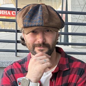 May include: A brown corduroy newsboy cap with blue and brown patterned panels, worn with a red and black buffalo plaid button-down shirt over a white t-shirt, featuring a white digital watch with a ribbed band and a silver ring on the finger.