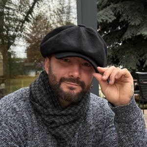 以下が含まれることがあります： 構造化されたクラウンと短く硬いブリッジを備えた黒いニュースボーイキャップ。キャップは中央にボタンのディテールがあるクラシックなマルチパネルデザインで、滑らかで均一な質感の無地の黒い生地で作られています。