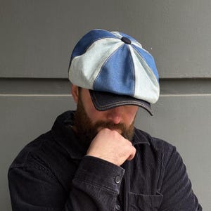 May include: A denim newsboy cap with a patchwork design in shades of blue and light blue. The cap has a dark blue brim and a button on top. The person in the image is wearing a dark blue corduroy shirt.