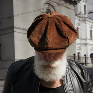 Puede incluir: Gorra de estilo newsboy de pana marrón. La gorra tiene una corona redondeada y una visera corta y rígida. La gorra es de color marrón cálido y parece estar hecha de una tela suave y texturizada. La persona lleva una larga barba blanca.