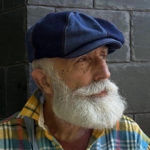 Puede incluir: Una gorra plana de mezclilla azul oscuro con una construcción de múltiples paneles y costuras visibles de color cobre a lo largo de las uniones. La gorra tiene una corona redondeada y una visera corta y rígida hecha de tela de algodón.