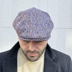 Puede incluir: Gorra de estilo newsboy con estampado de espiga en azul, marrón y blanco. La gorra tiene una corona redondeada y una pequeña visera rígida. Se lleva con una chaqueta oscura y un jersey de cuello alto color crema.
