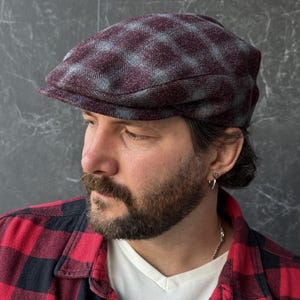 以下が含まれることがあります： ダークバーガンディとグレーのチェック柄のフラットキャップ。このキャップは、構造化された丸みを帯びたクラウンと、短く硬いブリムを備えています。生地はウールのような質感で、表面全体に一貫したチェック柄のデザインが施されています。