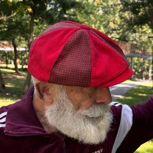 May include: A multi-paneled newsboy cap featuring alternating sections of bright red corduroy fabric and a dark brown and black micro-check patterned fabric. The cap has a rounded crown with a central button and a short, stiffened brim at the front.