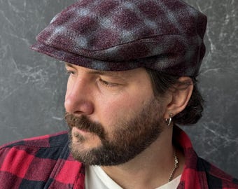 May include: A flat cap featuring a dark burgundy and grey plaid pattern. The cap has a structured, rounded crown and a short, stiff brim. The fabric has a textured, wool-like appearance with a consistent checkered design across the entire surface.