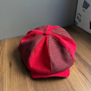 Peut inclure: Une casquette gavroche composée de panneaux alternés en velours côtelé rouge vif et en tissu à micro-carreaux marron et rouge. Le chapeau présente une forme arrondie à plusieurs panneaux avec une visière courte et rigide à l'avant.