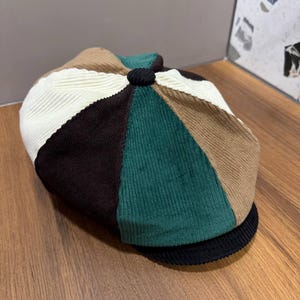 May include: A multi-colored corduroy newsboy cap featuring a patchwork design of cream, dark brown, tan, and forest green panels. The cap has a small black fabric-covered button at the center top and a short, curved black corduroy brim.