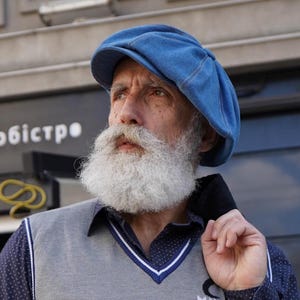 May include: A blue denim newsboy cap. The cap is worn by a person with a long white beard, a gray sweater vest, and a blue patterned shirt. The person is looking up and to the right.
