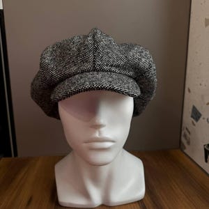 May include: A black and white tweed newsboy cap with a rounded crown and a short, stiff brim. The hat is displayed on a white mannequin head. The cap's fabric has a subtle, textured pattern.