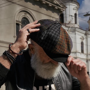May include: A multi-colored tweed newsboy cap with a black brim. The cap features a patchwork design with houndstooth and plaid patterns in shades of brown, gray, and purple. The cap is worn by a person with a white beard.